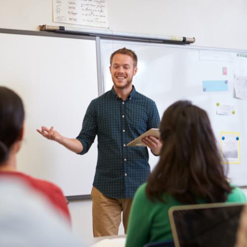 Junger Lehrer steht lächelnd mit einem Tablet vor der Klasse. | make & mint - mintLab Personal