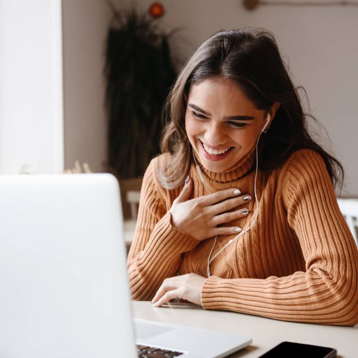 Attraktive junge Frau sitzt lächelnd vor ihrem Laptop und facetimed mit Kopfhörern im Ohr. | make & mint - Ablauf Förderung Schritt 3